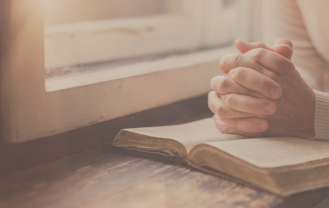 Hands in prayer on an open Bible by a window, representing faith and reflection at Grace Online Church Prayer Request page