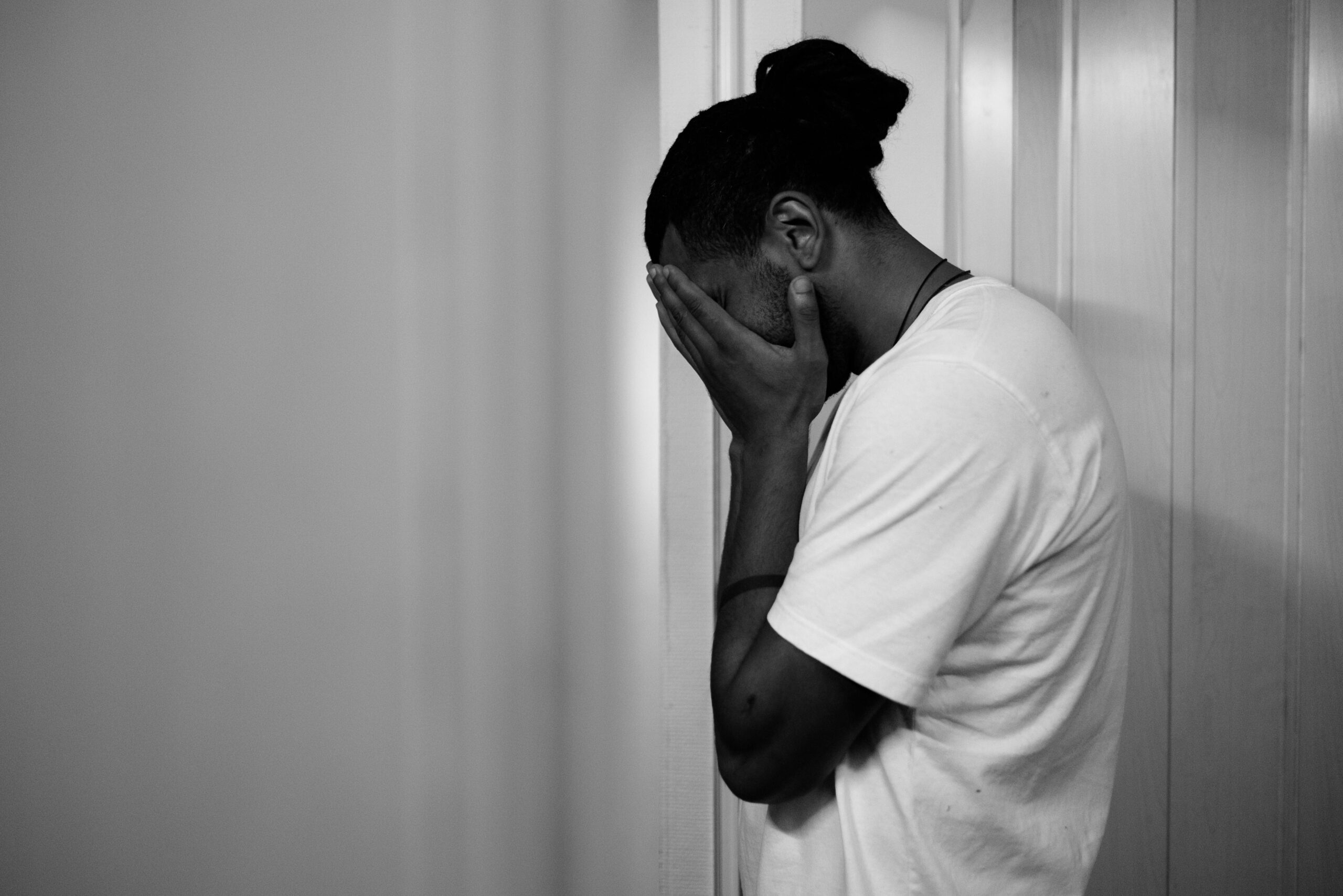 Grace Online Church presents a black and white photograph of a man standing alone, leaning against a wall with his hands covering his face. This powerful image symbolizes emotional struggle, signs of depression, and the importance of mental health awareness in the Black community